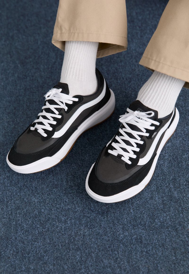 Scarpe da ginnastica nere e grigie con lacci bianchi, materiale testurizzato e suola in gomma, posizionate su una moquette blu, abbinate a calzini bianchi a coste.
