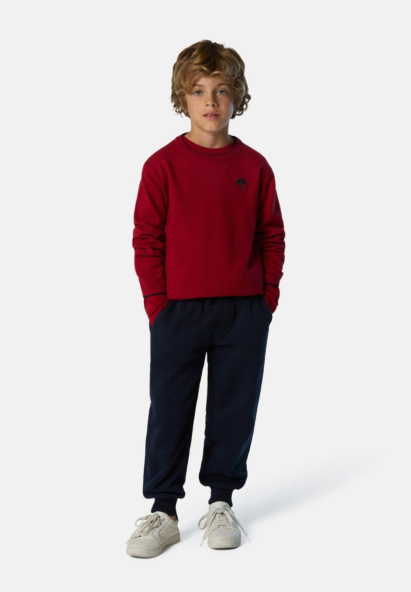Sudadera roja con cuello redondo y logo negro, combinada con pantalones de chándal azul marino y zapatillas blancas.