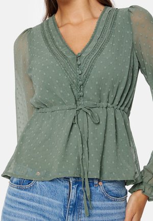 Kobieta ubrana w przezroczystą bluzkę w kolorze szałwi zielonej z teksturowanymi kropkami, koronkowym wykończeniem, guzikami obciągniętymi tkaniną, paskiem w talii i długimi rękawami, zestawioną z niebieskimi dżinsami.