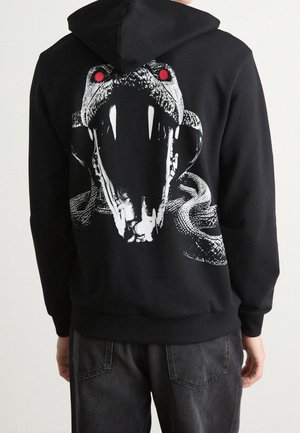 Sweat à capuche noir avec un grand motif de serpent blanc dans le dos, avec une bouche ouverte, des crocs visibles et des yeux rouges.
