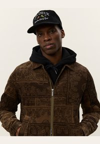 Chaqueta de pana marrón con un patrón intrincado, cierre de cremallera y dos bolsillos laterales, llevada sobre una sudadera negra con capucha y combinada con una gorra de marca.