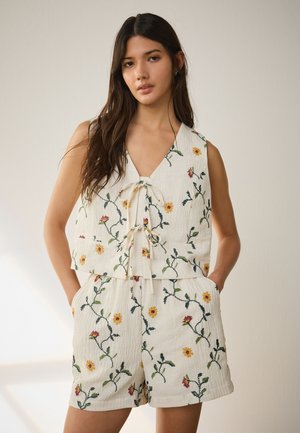 Giovane donna che indossa un top bianco senza maniche e pantaloncini con ricamo floreale giallo, in piedi con le mani nelle tasche davanti a uno sfondo neutro.
