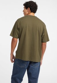 Tricou din bumbac verde măsliniu, cu croială lejeră, mâneci scurte, guler rotund și fără grafică vizibilă. Purtați cu blugi albastri din denim.
