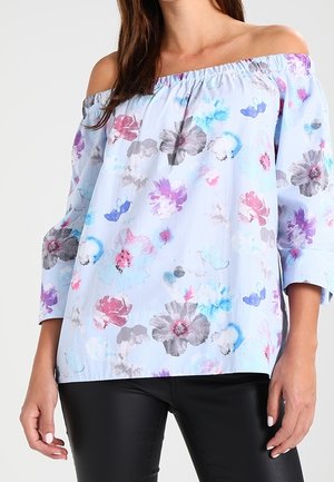 Femme portant un chemisier bleu clair à épaules dénudées avec des imprimés floraux multicolores et un pantalon noir, debout avec les bras détendus le long du corps.
