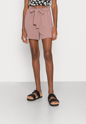 Femme portant un short taille paperbag mauve avec ceinture nouée et des sandales noires, debout devant un fond uni clair.