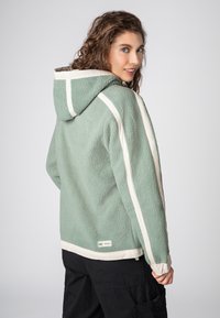 Groen fleecejack met crèmekleurige accenten, capuchon, gestructureerde stof en verstelbare manchetten, gezien vanuit een achteraanzicht. Zwarte cargobroek zichtbaar.