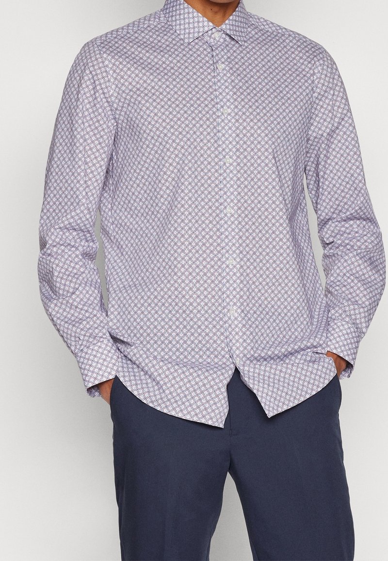Homme portant une chemise à manches longues boutonnée avec un motif géométrique violet clair et un pantalon bleu foncé, mains dans les poches.