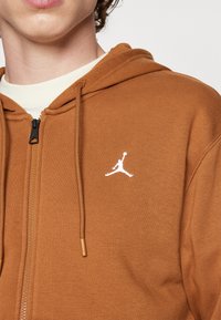 Personne portant un sweat à capuche marron zippé avec un logo Jumpman blanc sur la poitrine et une chemise blanche en dessous.