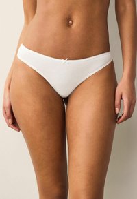 String en coton blanc avec une texture lisse, présentant un petit nœud à l'avant et des bords festonnés délicats.