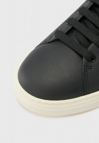 Black leather sneaker with textured surface, rounded toe, white rubber sole, and black laces. The design features minimal stitching details.