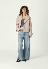MLADA ŽENSKA S KOVRČASTO KOSEMU OBLEČENA V BEŽ KRAČNO JAKNO, MODREM KRAČNEM TOPU, ŠIROKOHLAHAVE LAHKO MODRE JEANS IN ČRNIH ŠPICAH ČEVLJIH, STOJI.