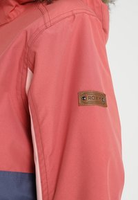 Różowa wodoodporna kurtka z brązową skórzaną łatą z logo Roxy na rękawie, o gładkiej fakturze i kontrastujących elementach wzorniczych.