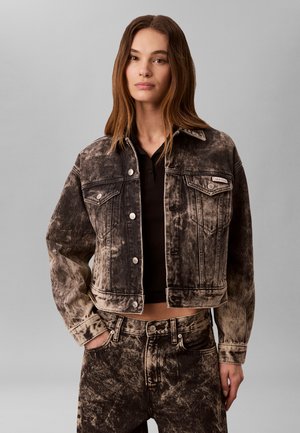 Junge Frau mit langen braunen Haaren, die eine schwarze, kurz geschnittene, gewaschene Jeansjacke und passende hoch taillierte, gewaschene Jeanshosen trägt.