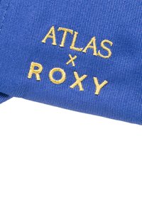 Blue fabric with a textured weave featuring yellow embroidered text "ATLAS x ROXY". The design focuses on bold, modern typography.