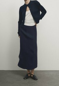 Marineblauwe gebreide cardigan met ronde kraag, gecombineerd met een lange marineblauwe rok. Eenvoudig wit hemdje eronder. Zwarte platte schoenen met enkelbandjes.