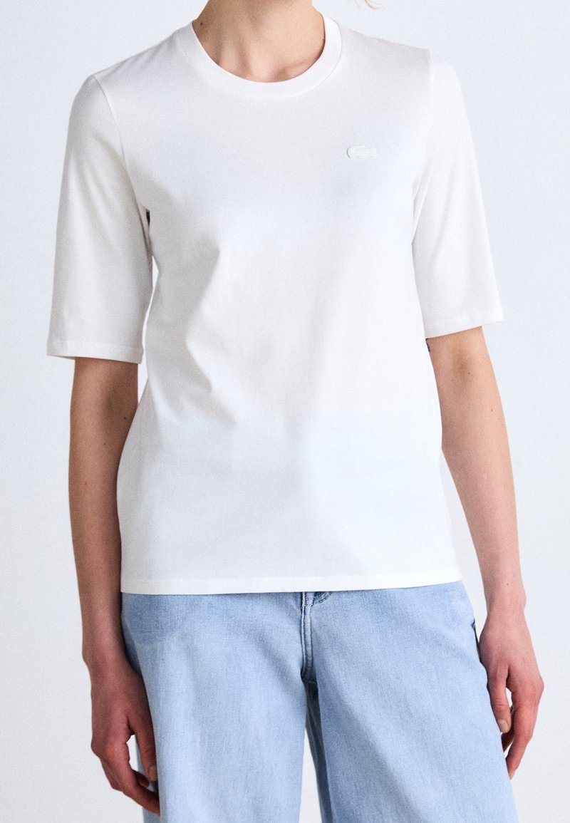 White cotton t-shirt with short sleeves, crew neckline, and a small tonal logo on the chest. Paired with light blue denim jeans.