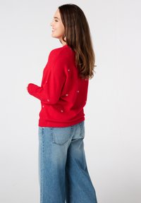 Pull rouge en maille légère, avec de petits motifs en cœur argentés, associé à un jean clair à jambes larges.