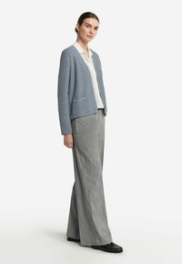 Helles blaues, strukturiertes Cardigan mit zwei vorderen Taschen, getragen über einem weißen Hemd und grauen gestreiften weiten Hosen, kombiniert mit schwarzen Schuhen.