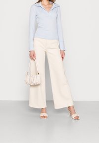 Chemise polo à manches longues bleu clair, large pantalon beige, sandales à brides blanches, et sac à main crème froncé. Texture lisse, coupe décontractée.