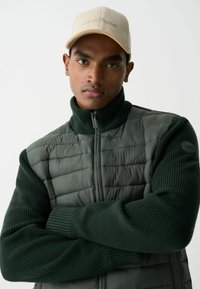 Paminkštintas žalias švarkas su aukštu apykakle ir plyšiais megztose rankovėse, derinamas su smėlio spalvos velcro kepure. Papuoštas užtrauktuku ir logotipo detale.