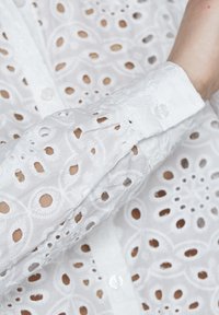 Hvid broderet bluse med uds�øgte udskæringer, en knaplukning og en struktureret manchet med en enkelt knapdetalje.