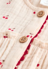 Tissu beige clair avec des plis, orné de motifs de cerises en rouge et marron. Présente des boutons en bois et une bordure à pompons rouges le long de l'ouverture.