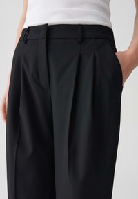Zwarte geplooide broek van gladde stof met een tailored fit, voorzien van zijzakken en een platte tailleband voor een nette afwerking.