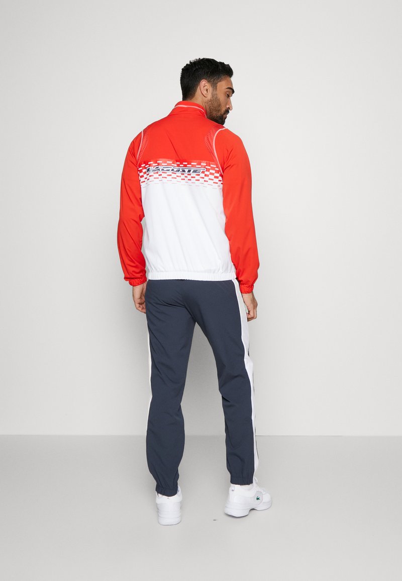 Giacca a vento rossa e bianca con motivo a scacchi sul retro, polsini elasticizzati. Abbinata a pantaloni da jogging blu navy con strisce bianche sui lati. Sneakers bianche.