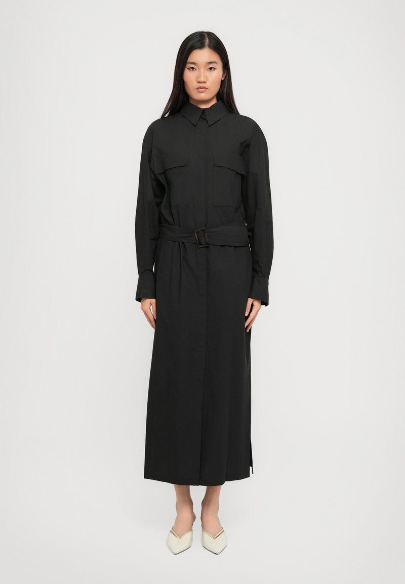 Black long-sleeve shirt dress with collar, front pockets, belted waist, and side slits, made from lightweight fabric; paired with cream pointed shoes.