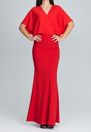 Femme portant une robe rouge longue jusqu'au sol avec un profond décolleté en V et des manches larges, les mains posées sur les hanches, debout devant un fond uni.