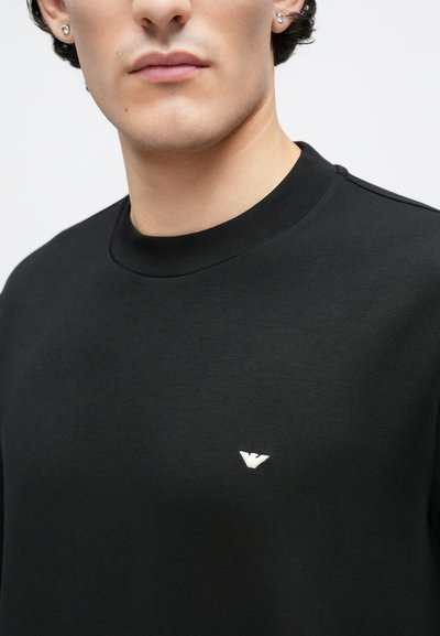 Čierna mikina z hladkej tkaniny, s okrúhlym výstrihom a malým bielym logom na prsiach. Jednoduchý a minimalistický dizajn.