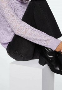 Maglione lavorato a maglia di un delicato viola chiaro con motivo in pizzo, abbinato a jeans in denim neri a vita alta e stivaletti neri con tacco, seduto su un pedestal bianco.