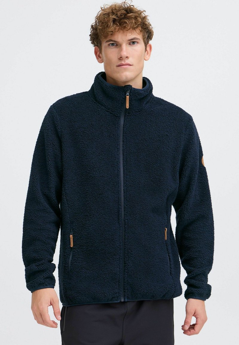 Marineblaue Fleecejacke mit hohem Kragen, durchgehendem Reißverschluss und Seitentaschen. Verfügt über einen strukturierten Stoff und einen Reißverschluss-Zipper mit Holzakzent.