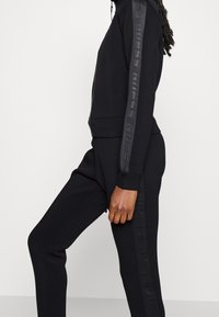 Seitenansicht einer Person, die eine schwarze Jacke mit Reißverschluss und passende Hose trägt, beide mit tonalen Logosstreifen an Ärmeln und Beinen.