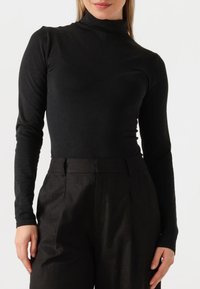 Schwarzer Rollkragenpullover im figurbetonten Design mit langen Ärmeln, aus weichem Stoff, getragen zu hochgeschnittenen dunklen Hosen. Sanfte Textur, minimale Details.
