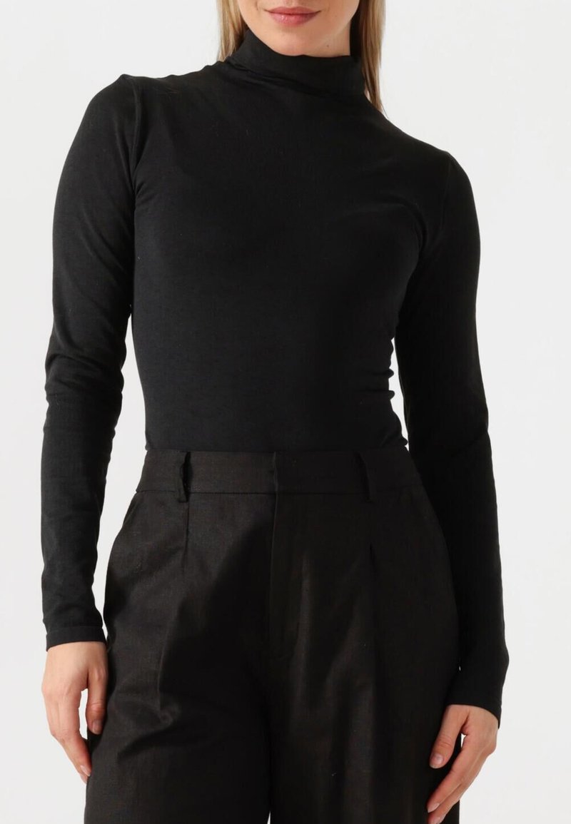 Schwarzer Rollkragenpullover im figurbetonten Design mit langen Ärmeln, aus weichem Stoff, getragen zu hochgeschnittenen dunklen Hosen. Sanfte Textur, minimale Details.