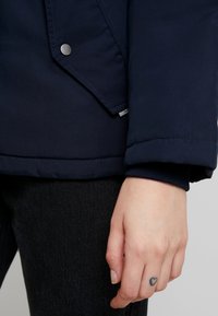 Main avec un tatouage en forme de cœur sur le doigt à côté du poignet de la veste bleu marine et de la poche à bouton pression, au-dessus d'un pantalon noir.