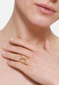 Goldener Metallring mit zwei miteinander verflochtenen ovalen Formen. Glatte Oberfläche, schlankes Design, getragen an einer Hand, die gegen einen Hals ruht.
