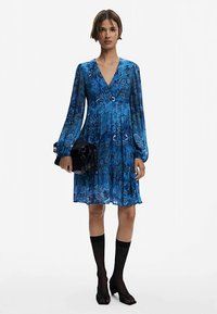 Blaues Blumenkleid mit langen Ärmeln und V-Ausschnitt, strukturierter Stoff, plissiertes Design, kombiniert mit einer schwarzen Handtasche und kniehohen schwarzen Socken.