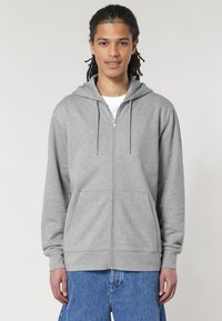 Felpa grigia con zip realizzata in un tessuto morbido, dotata di tasca frontale, lacci sul cappuccio e polsini a coste. Indossata sopra una maglietta bianca.