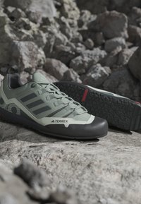 Ανοιχτό γκρι παπούτσια Adidas Terrex για μονοπάτια με μαύρες σόλες, εμφανιζόμενα σε βραχώδες έδαφος με ένα παπούτσι ξαπλωμένο στο πλάι που δείχνει το σχέδιο της σόλας.