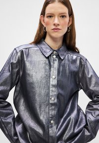 Camisa azul metálica com botão frontal, colarinho clássico e mangas longas. Apresenta uma textura suave e botões prateados.