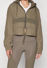 Chaqueta verde oliva recortada con cremallera en la parte delantera, capucha y cinturilla elástica. Presenta una banda ribeteada horizontal y mangas acampanadas.