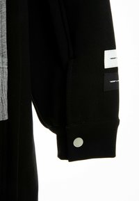 Manga de sudadera negra con un panel acanalado gris, que presenta tres etiquetas en blanco y negro, y un botón a presión plateado en el puño.