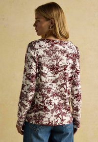 Dlhý rukáv v krémovej farbe s burgundským toile vzorom, ktorý obsahuje stromy, postavy a krajiny. Okrouhly výstrih a priliehavé rukávy.