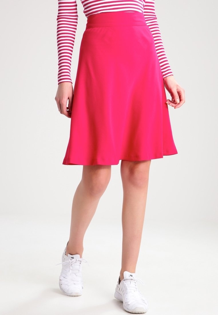 Femme portant une jupe rose vif longueur genou, un haut rayé rouge et blanc, et des baskets blanches, debout devant un fond blanc uni.