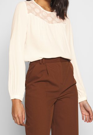 Kvinde iført en cremefarvet bluse med lange ærmer, gennemsigtig polkaprik-yoke og højttaljede brune skræddersyede bukser.