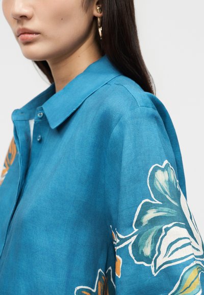 Γυναίκα που φοράει μπλε πουκάμισο με κουμπιά και γιακά, με μεγάλα λουλουδάτα σχέδια σε λευκό, πράσινο και πορτοκαλί στο μανίκι.