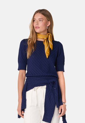 Pull en maille texturée bleu marine à manches courtes et bords festonnés, noué à la taille avec un tissu assorti. Écharpe à pois jaunes en accent.