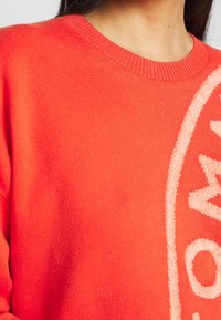 Gros plan d'une personne portant un pull en maille orange vif avec un logo circulaire blanc partiellement visible sur le côté droit.